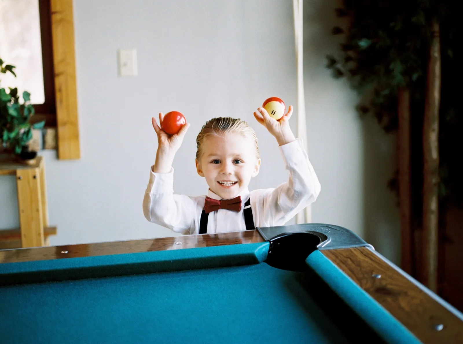 A young boy in a white shirt, bow tie, and burgundy suspenders holds two pool balls up near his face.