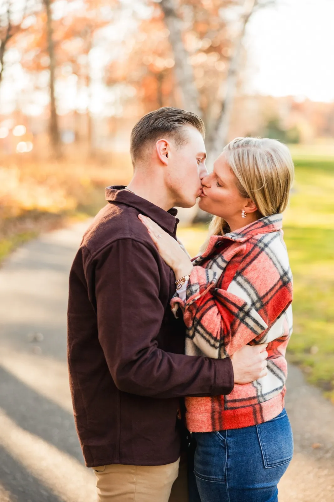 Tyler and Maddy kiss on an autumn path, her engagement ring visible against his dark shirt