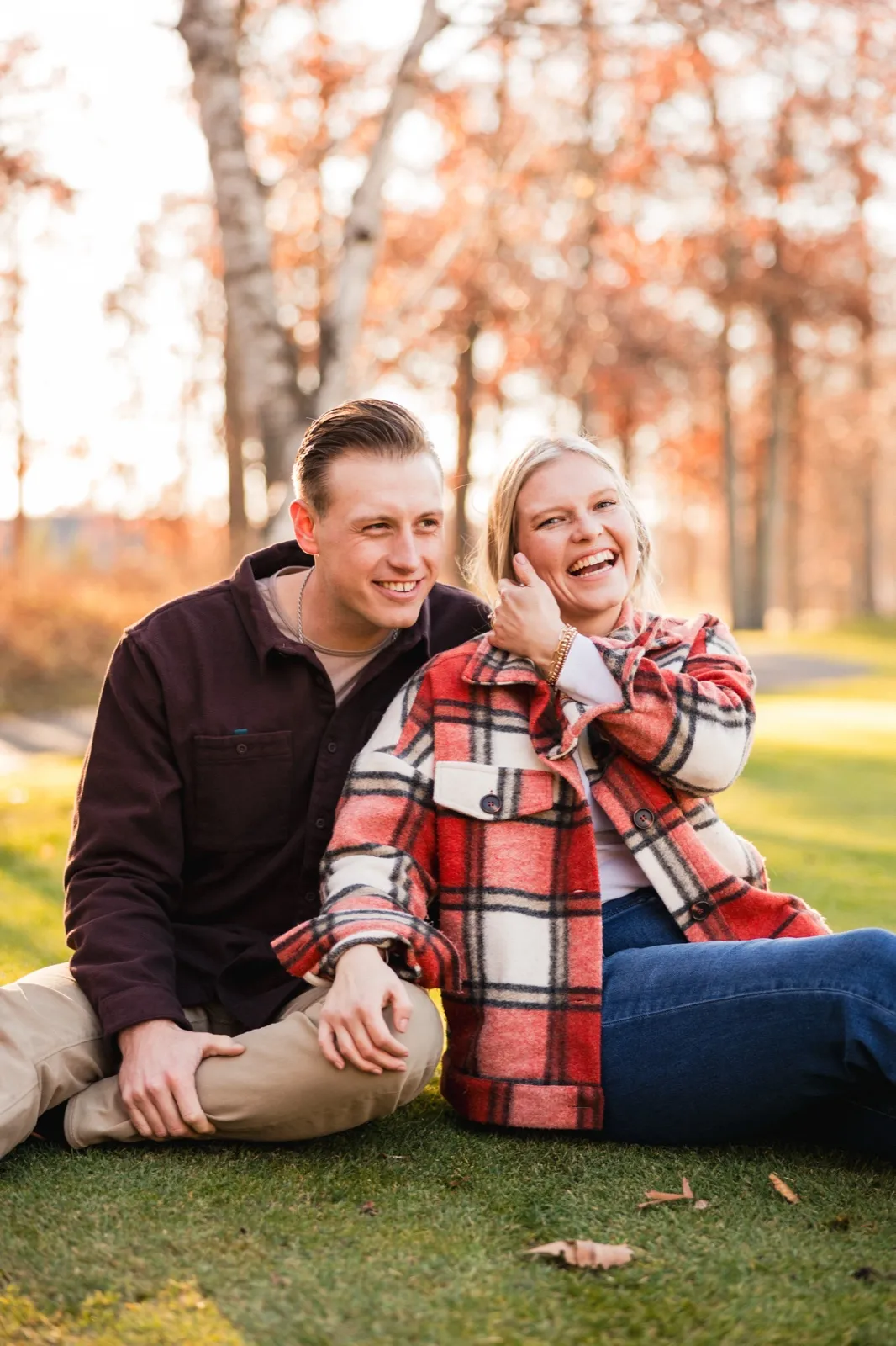 Maddy laughs as she and Tyler sit together on the grass, autumn trees glowing behind them — Tim Larsen Photography, Brainerd Lakes MN