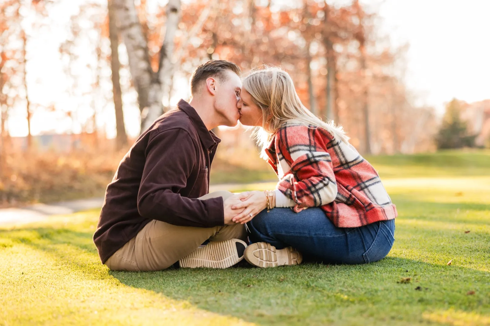 Maddy and Tyler kiss while sitting on sunlit grass, backlight coming through the trees — Tim Larsen Photography, Brainerd Lakes MN