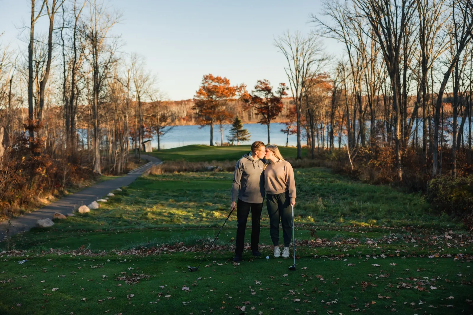 Maddy and Tyler kiss on a tee box, golf clubs in hand and a ball teed up, with the lake and autumn trees behind them — Tim Larsen Photography, Brainerd Lakes MN