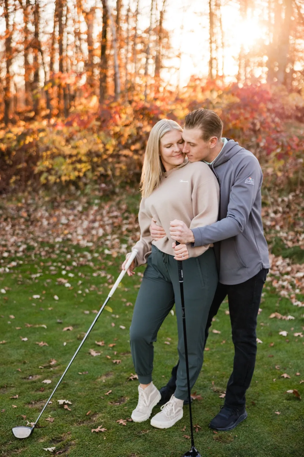 Tyler kisses Maddy as they stand on the course holding golf clubs, fall color behind them — Tim Larsen Photography, Brainerd Lakes MN