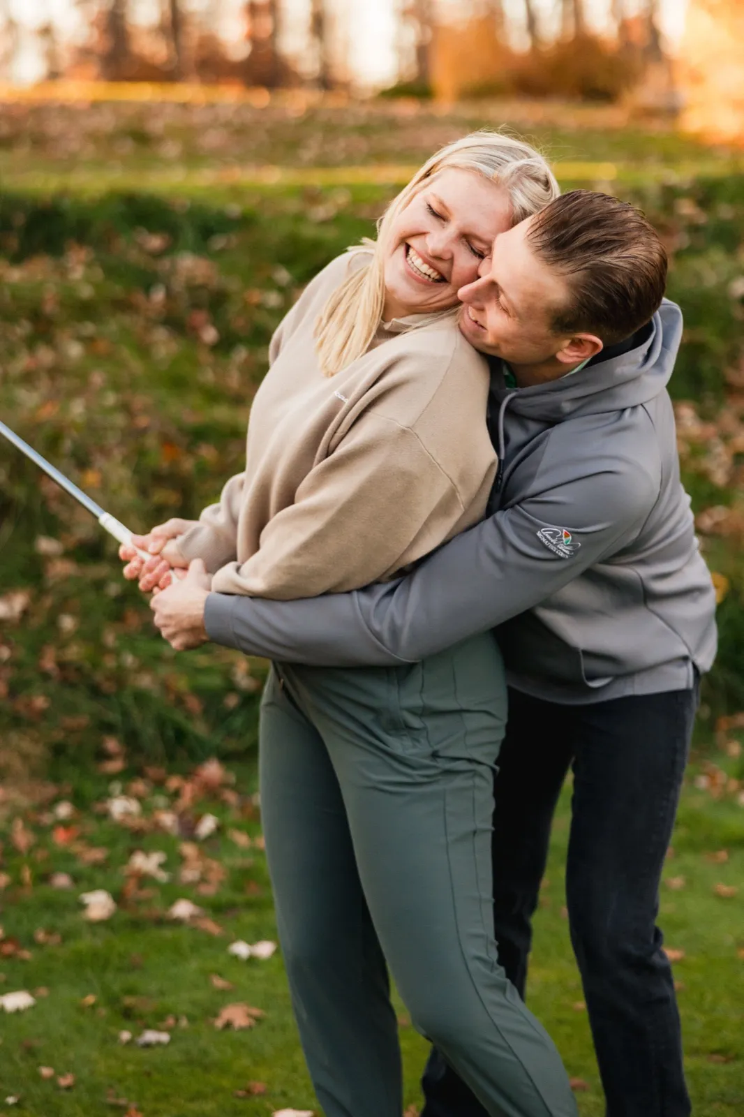 Tyler hugs Maddy from behind as she laughs, holding a golf club among fallen leaves — Tim Larsen Photography, Brainerd Lakes MN