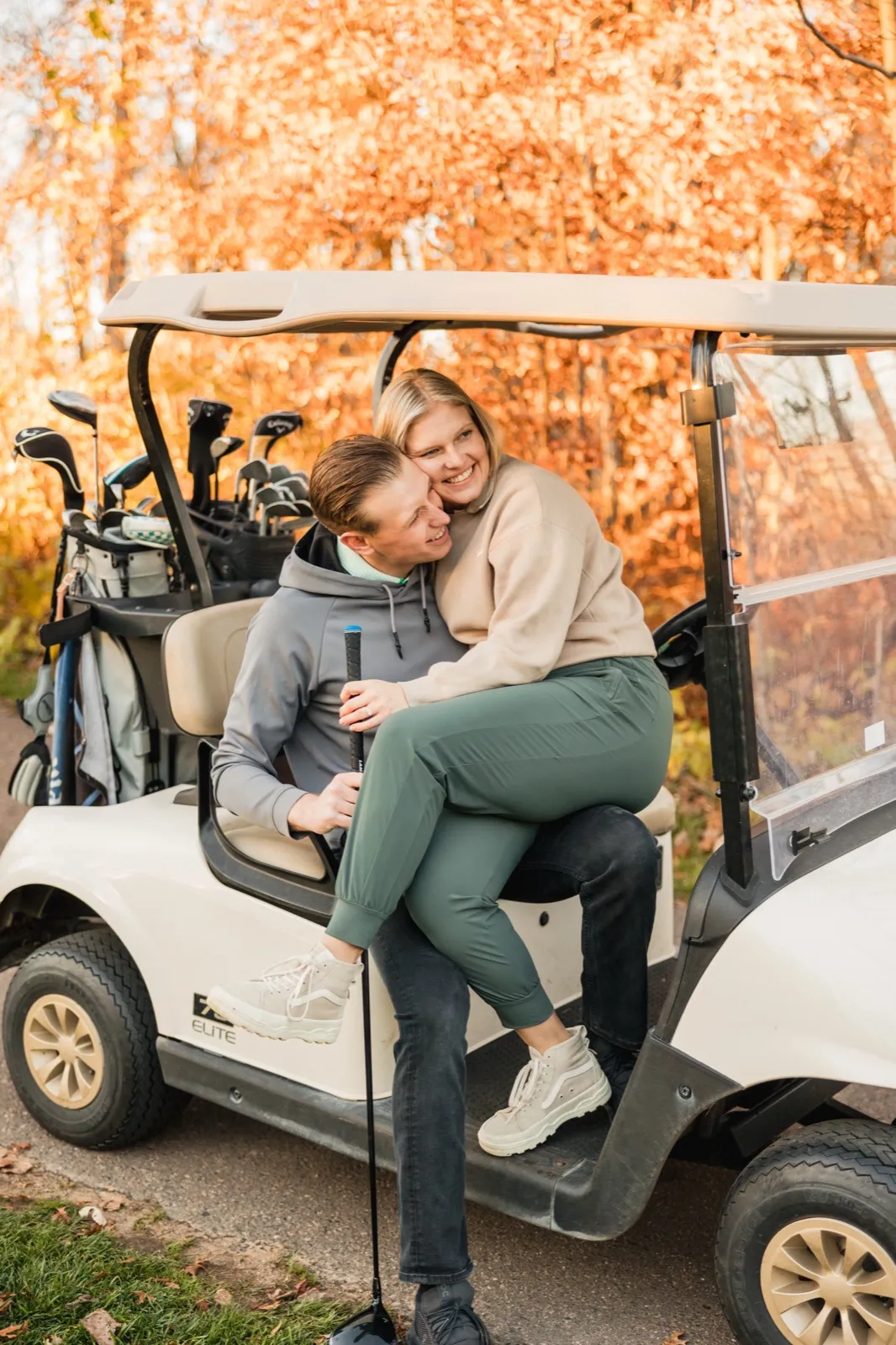 Maddy sits on Tyler's lap in a golf cart under orange autumn leaves at Crosswoods Golf Course