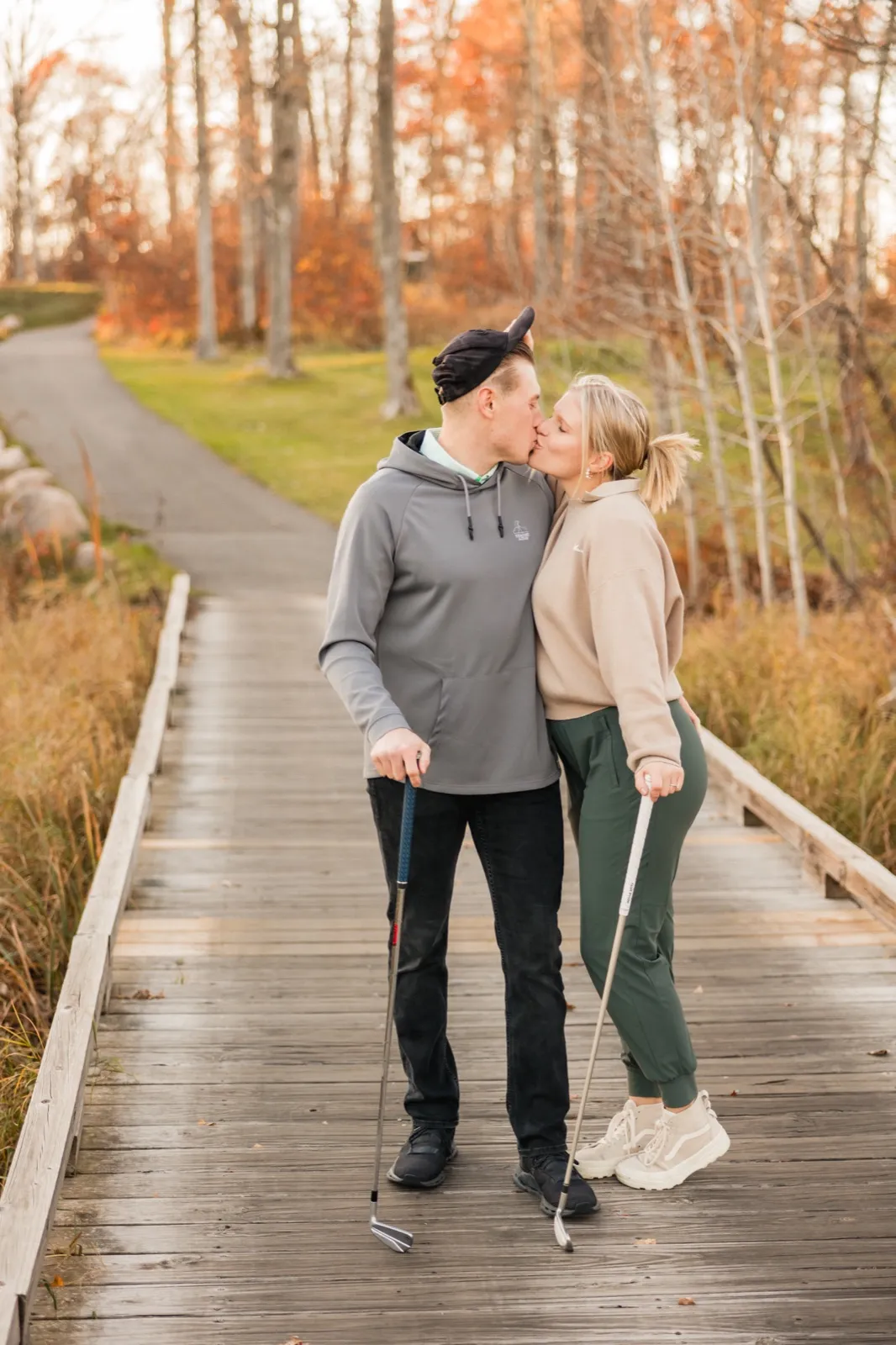 Maddy and Tyler kiss on a wooden boardwalk on the golf course, holding their clubs