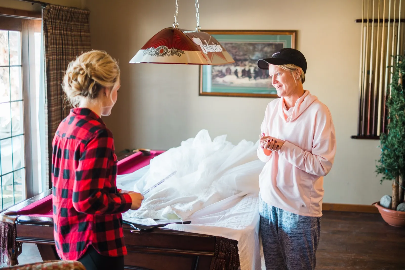 Morgan stands across a pool table from a woman in a pink hoodie, with her wedding dress spread between them.
