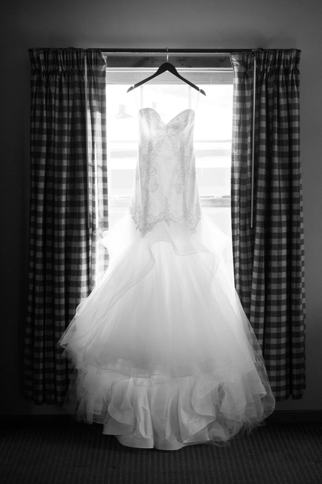 A black-and-white photograph of Morgan’s lace and tulle wedding gown hanging at a window framed by checked curtains.