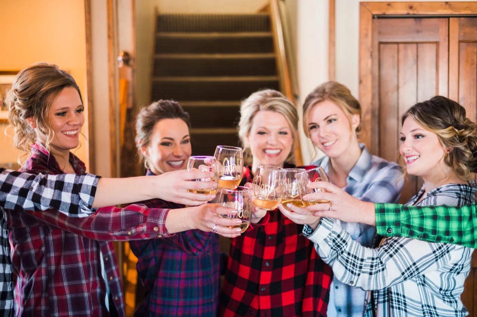 Morgan and her bridesmaids in different plaid shirts clink stemless glasses beside a wooden staircase.