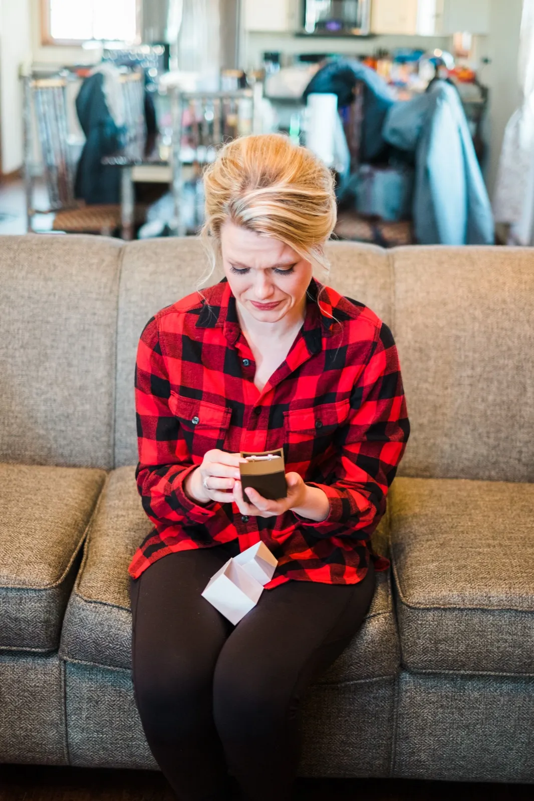 Morgan looks down at a small open gift box while sitting on a couch in her red plaid shirt.