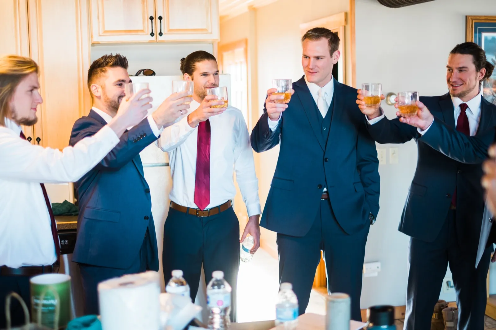 Cole and his groomsmen raise glasses in a sunlit kitchen, wearing blue waistcoats and white shirts.