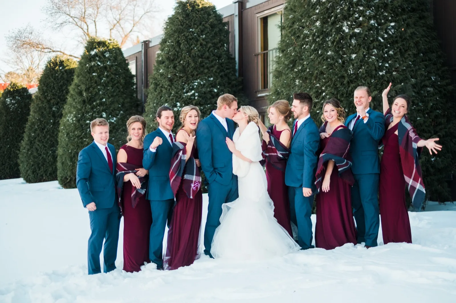 Morgan and Cole kiss in the snow while their wedding party in burgundy and blue laughs and cheers around them.