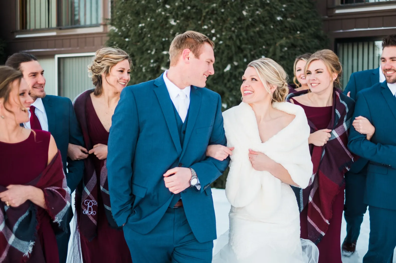 Morgan and Cole walk arm in arm through the snow with their wedding party, several bridesmaids wearing plaid wraps.