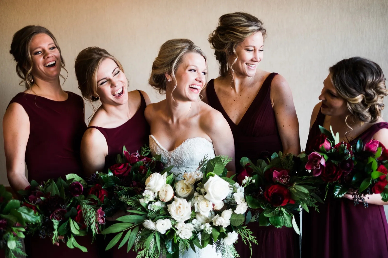 Morgan laughs with four bridesmaids in burgundy gowns as they hold their bouquets indoors.