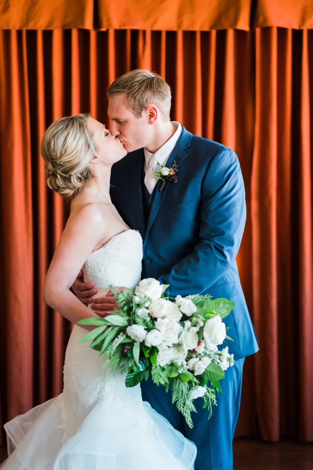 Morgan and Cole kiss in front of an orange curtain while she holds her white bouquet
