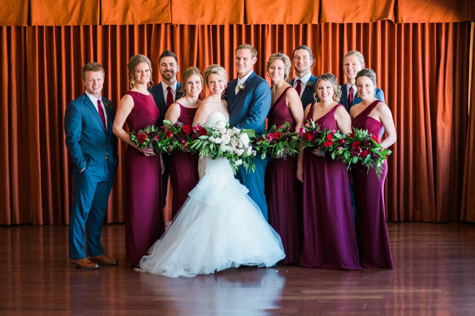 The wedding party stands before an orange curtain with bouquets held together across the front of the group.