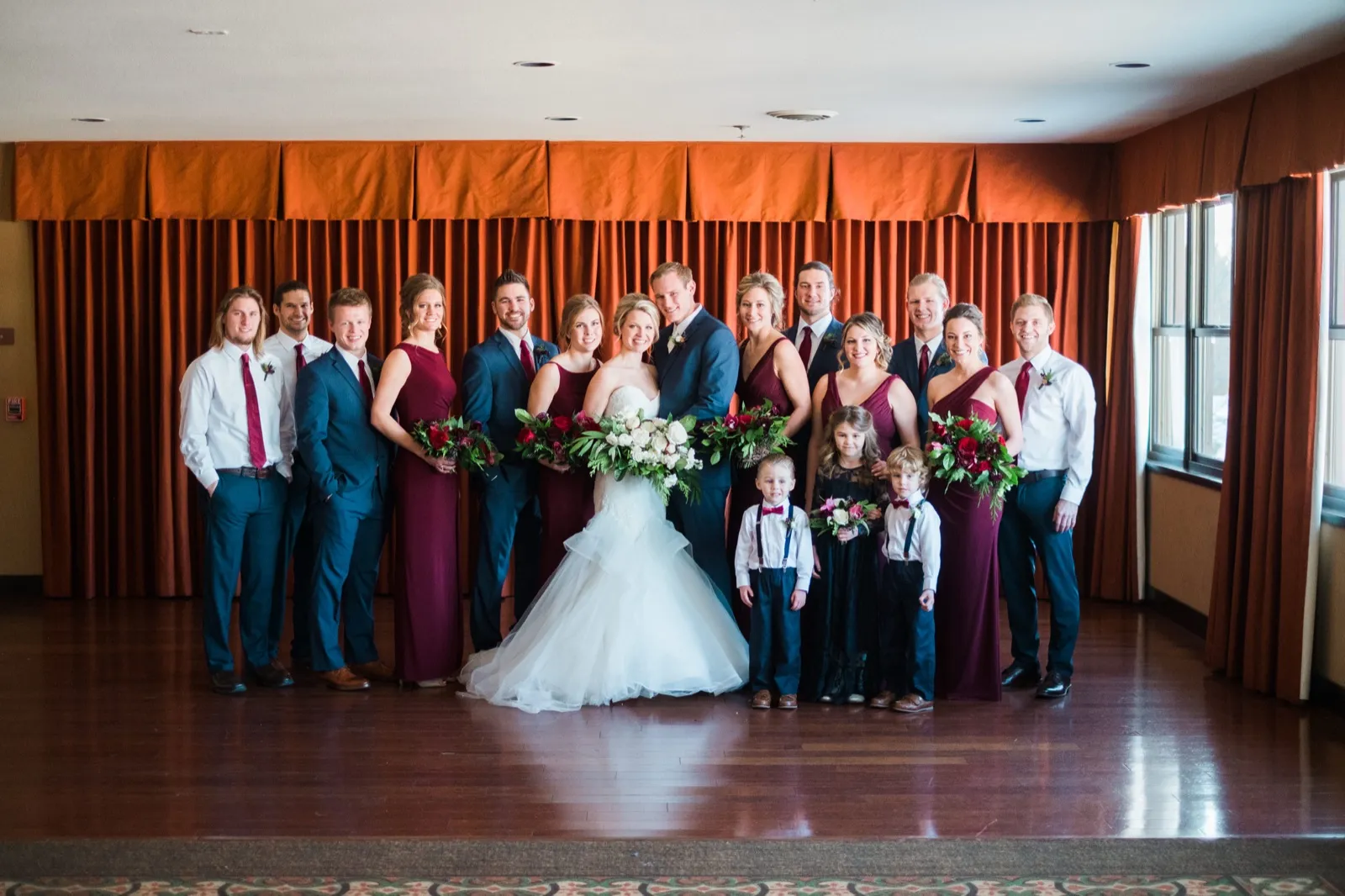 Morgan and Cole pose with their wedding party and young attendants in front of an orange curtain at Breezy Point Resort.