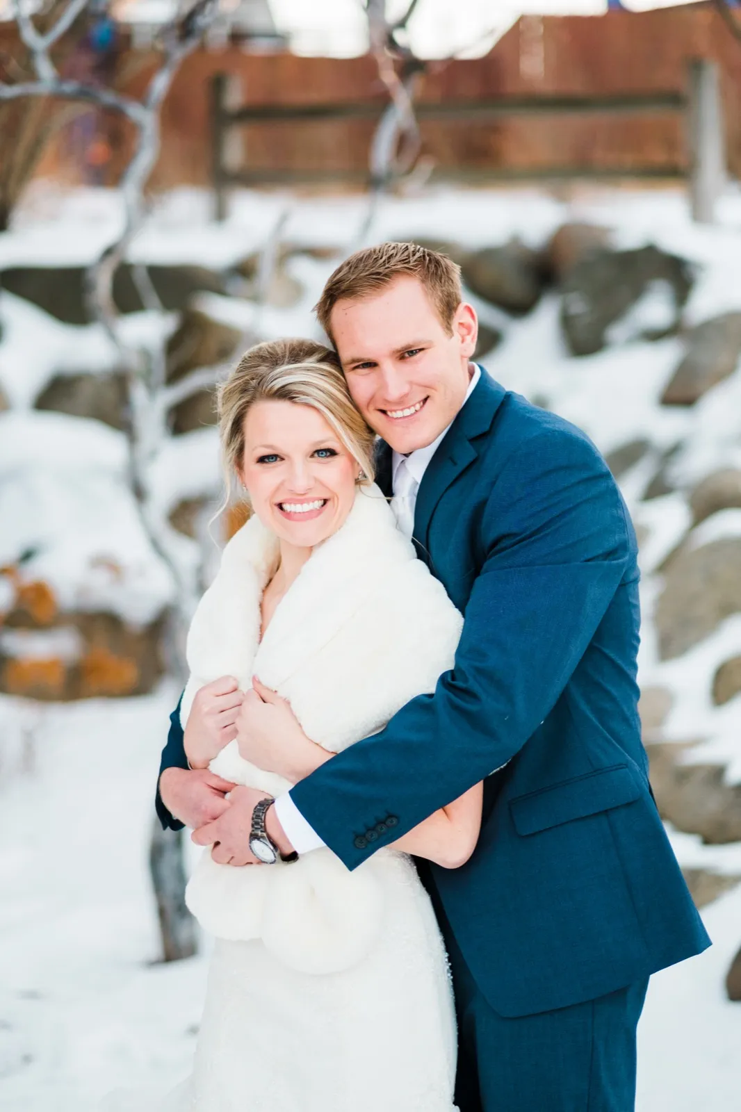 Morgan in her white wrap and Cole in his blue suit smile together in a snowy garden beside a wooden fence.