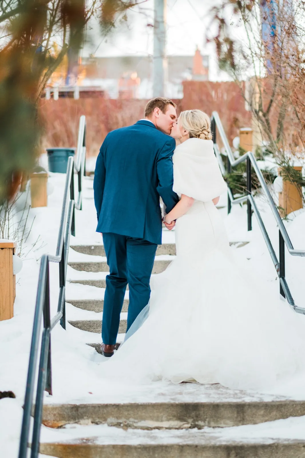 Morgan and Cole kiss on snowy stairs with green railings at Breezy Point Resort