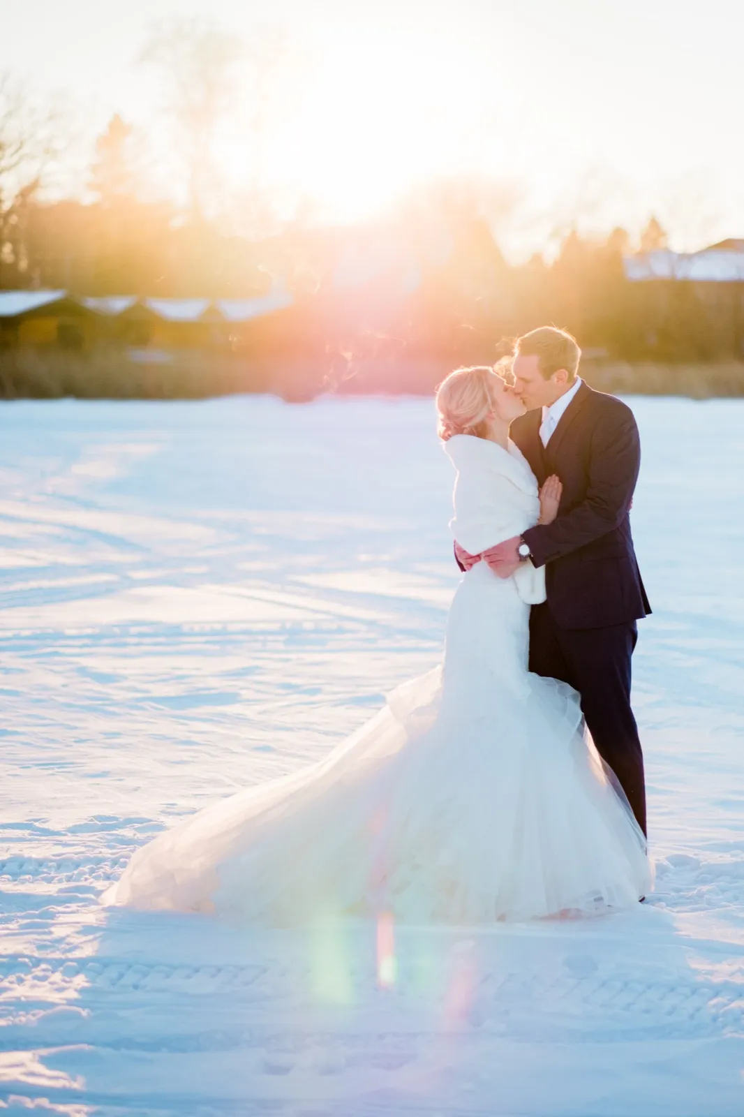 Morgan and Cole kiss on snow-covered Pelican Lake, with their breath and her white wrap lit by the low winter sun.