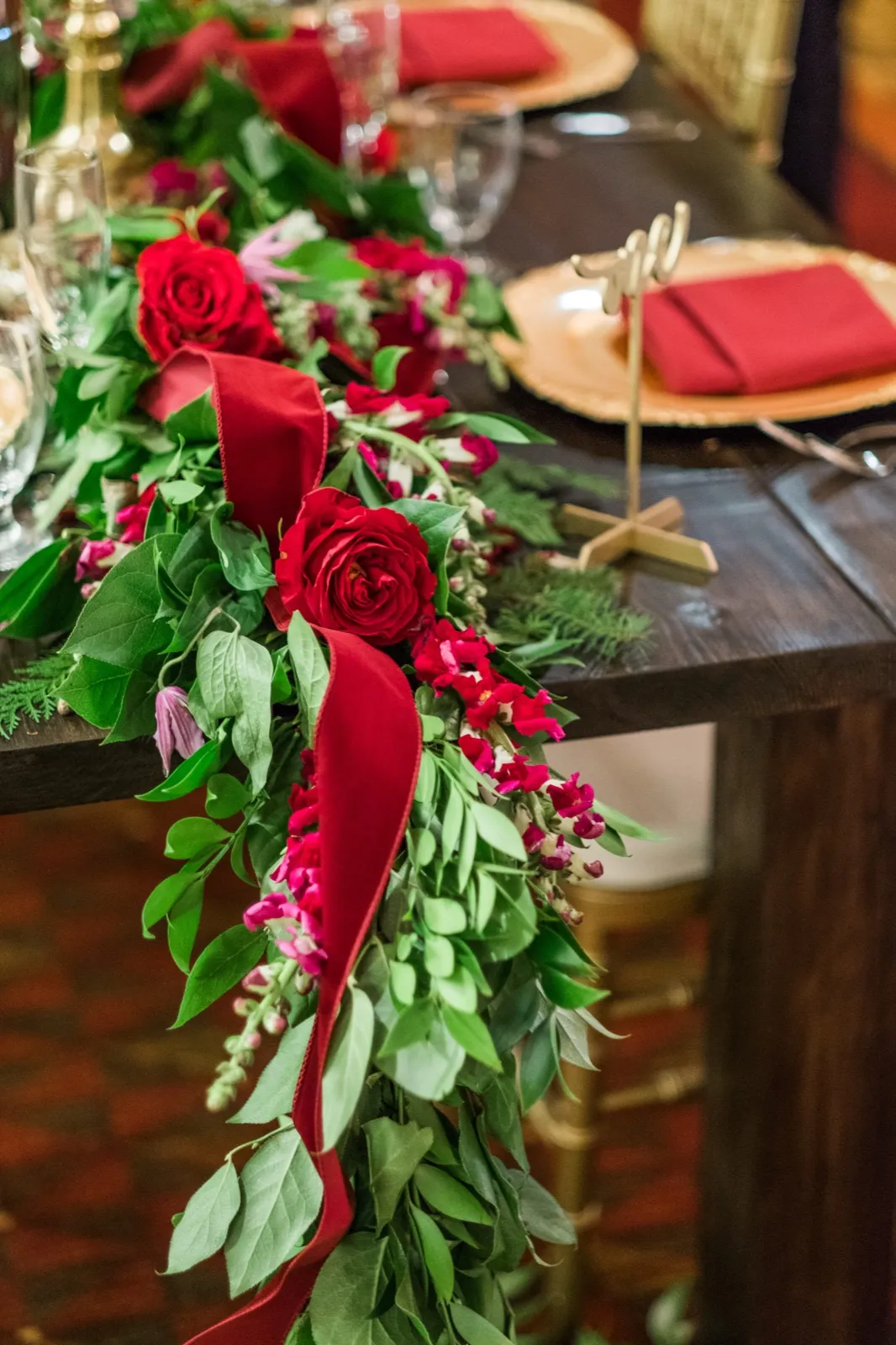 Red roses, greenery, and red ribbon trail over the edge of a reception table set with gold chargers.