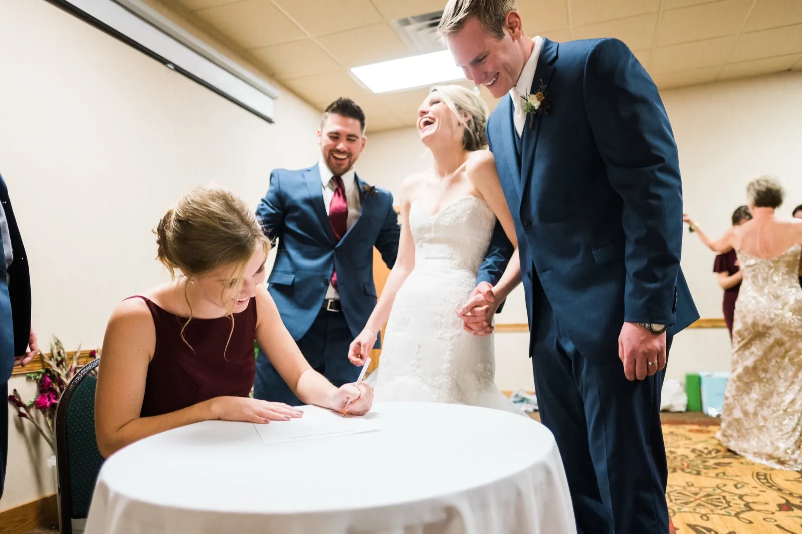 Morgan throws her head back laughing as a bridesmaid signs a document at a small table beside Cole.