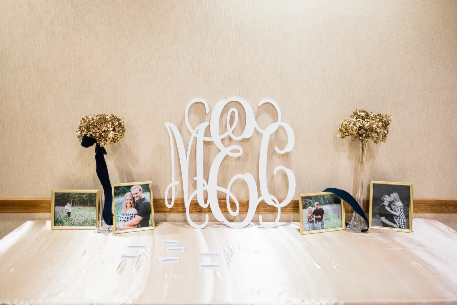 A white MCC monogram stands on a reception welcome table between framed photographs and tall gold arrangements.