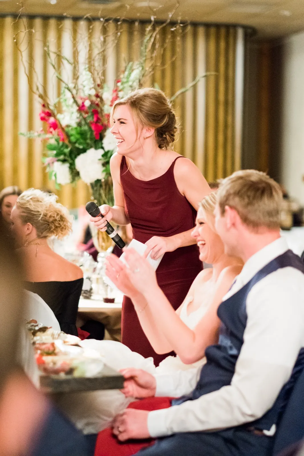 A bridesmaid in burgundy laughs while giving a toast as Morgan and Cole listen from their table.