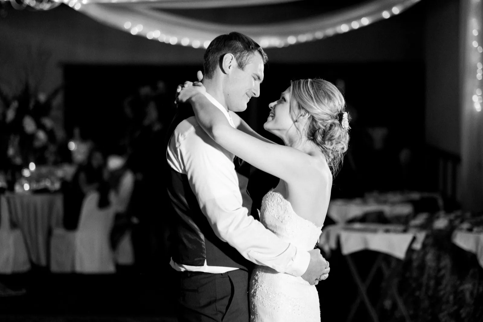 Morgan smiles up at Cole during their first dance beneath string lights, photographed in black and white.