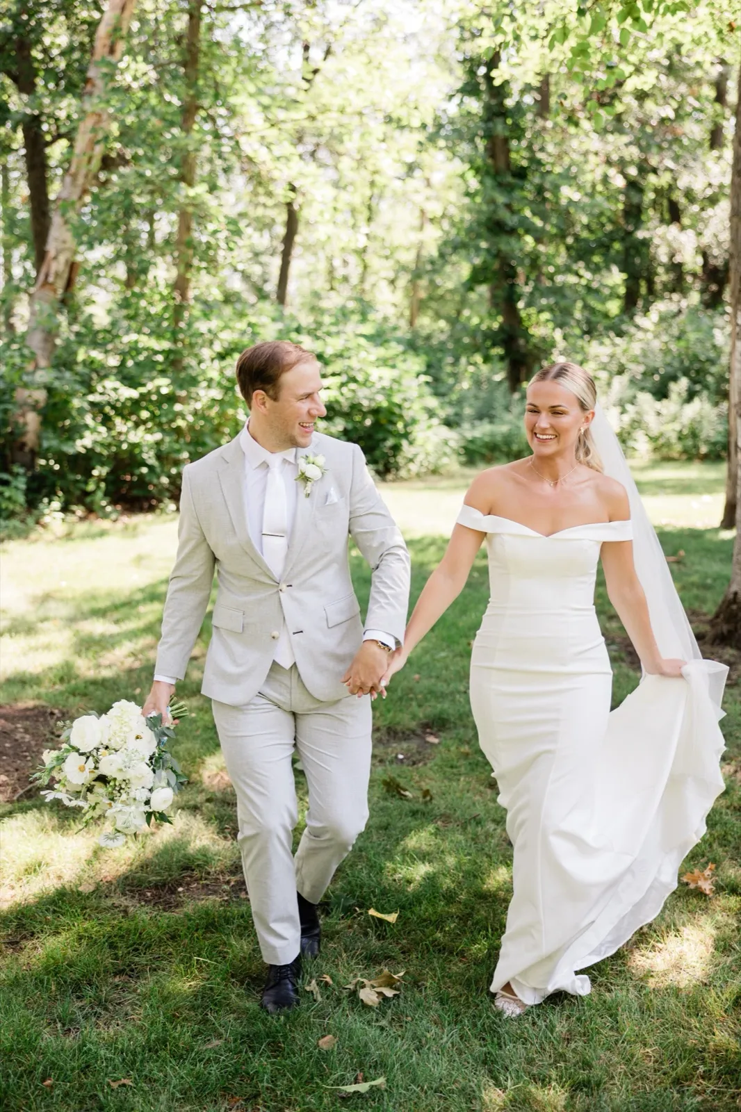 Josse holds the train of her gown as she and Alex walk hand in hand across a shaded lawn, both laughing — Tim Larsen Photography, Brainerd Lakes MN