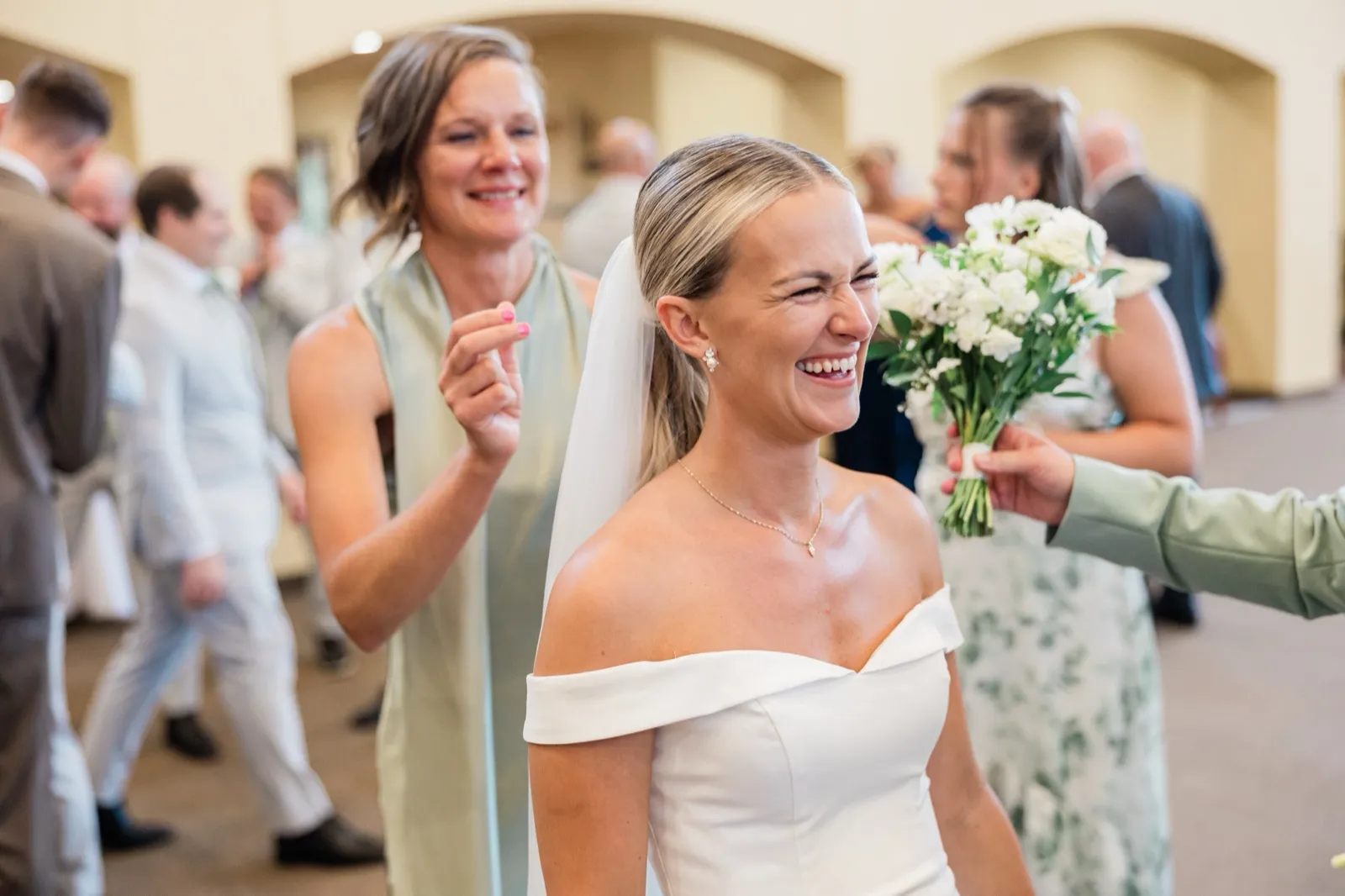 Josse laughs with her eyes squeezed shut as a bridesmaid hands her bouquet back to her in the church entryway — Tim Larsen Photography, Brainerd Lakes MN