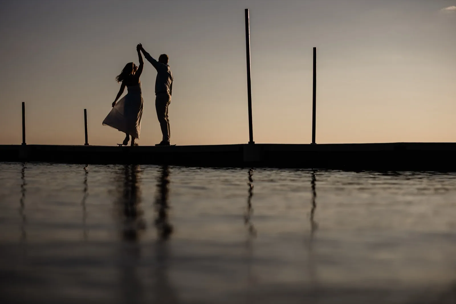 Adam twirls Olivia in silhouette on the dock at dusk, her dress flaring above the water's reflection — Tim Larsen Photography