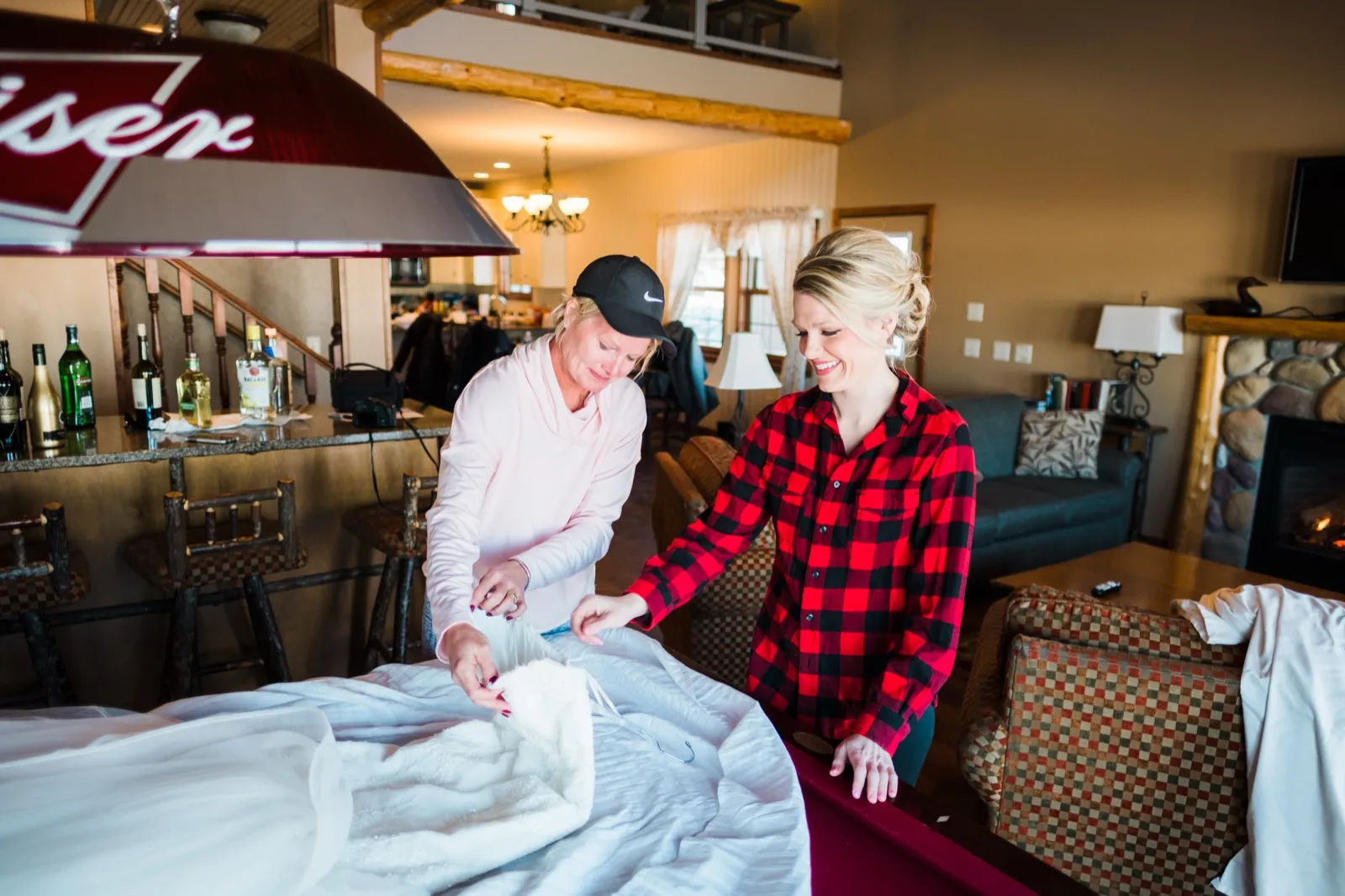 Morgan in a red plaid shirt smiles as a woman in a cap unpacks her wedding gown on a pool table beneath a Budweiser lamp.