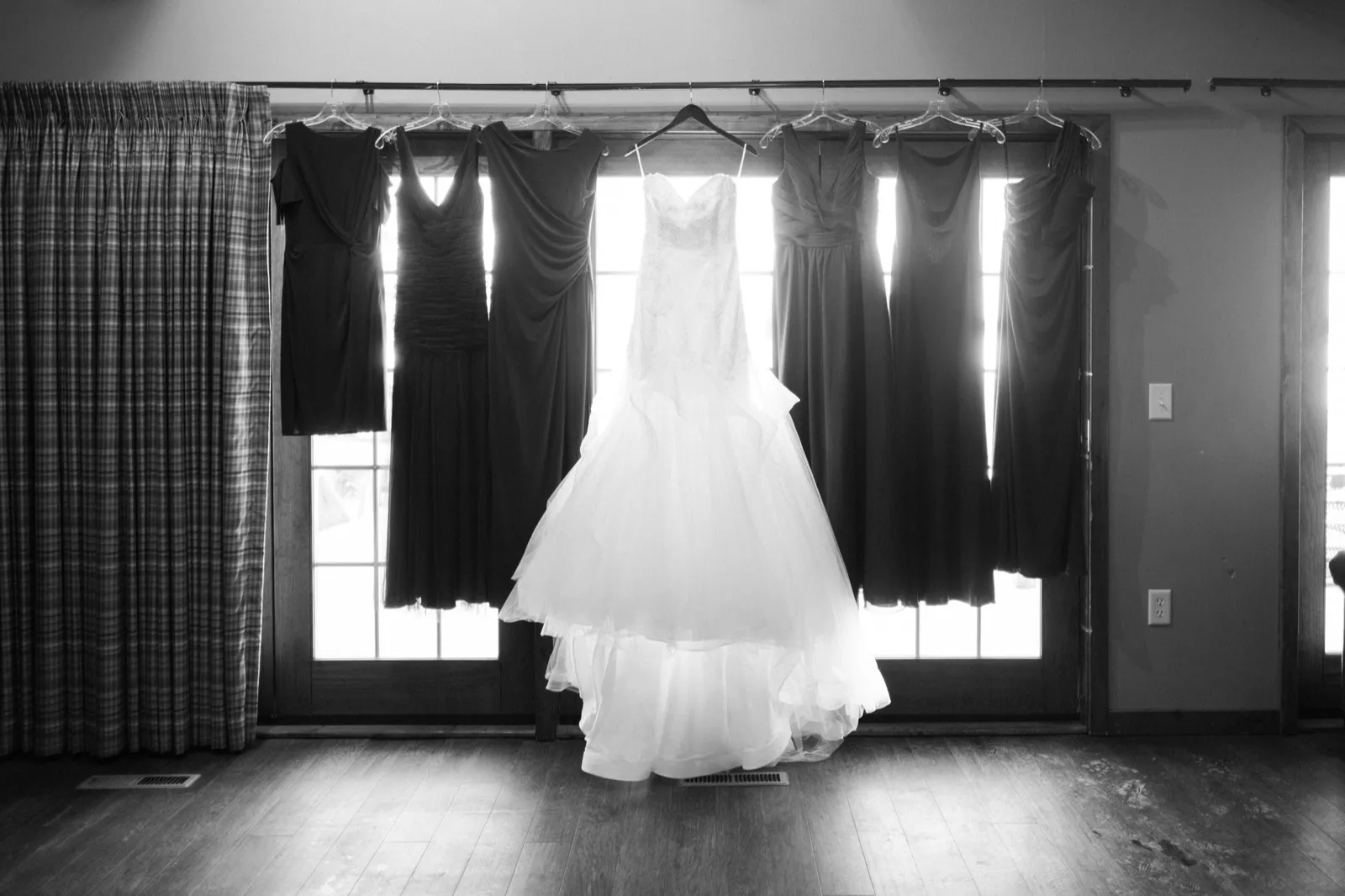 Morgan’s wedding gown hangs at a bright window between burgundy bridesmaid dresses, photographed in black and white.