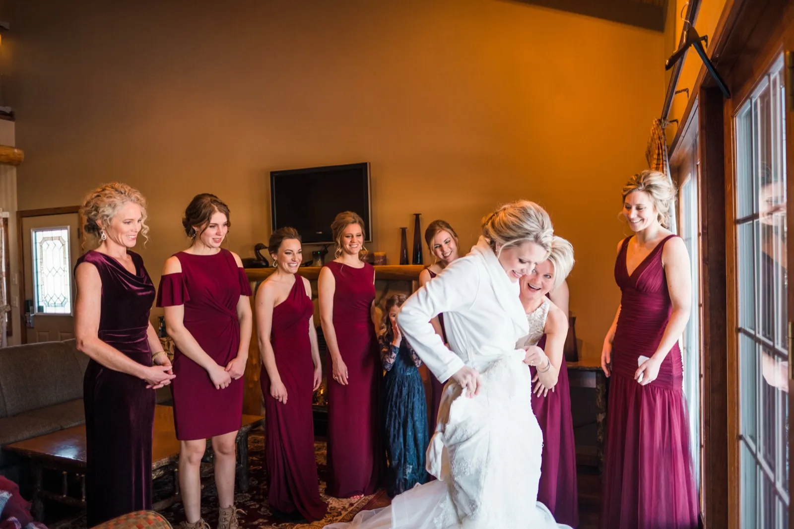 Morgan laughs as her mother in a silver dress helps fasten her wedding gown while bridesmaids watch.