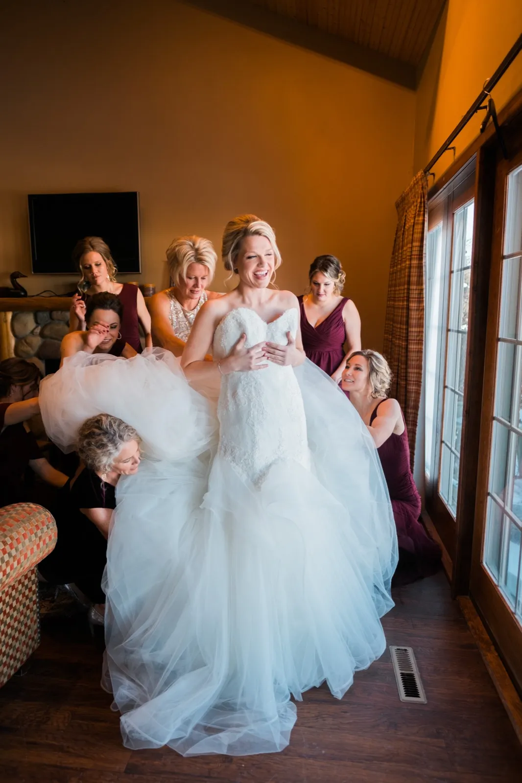 Morgan laughs while bridesmaids in burgundy help arrange her lace and tulle wedding gown.
