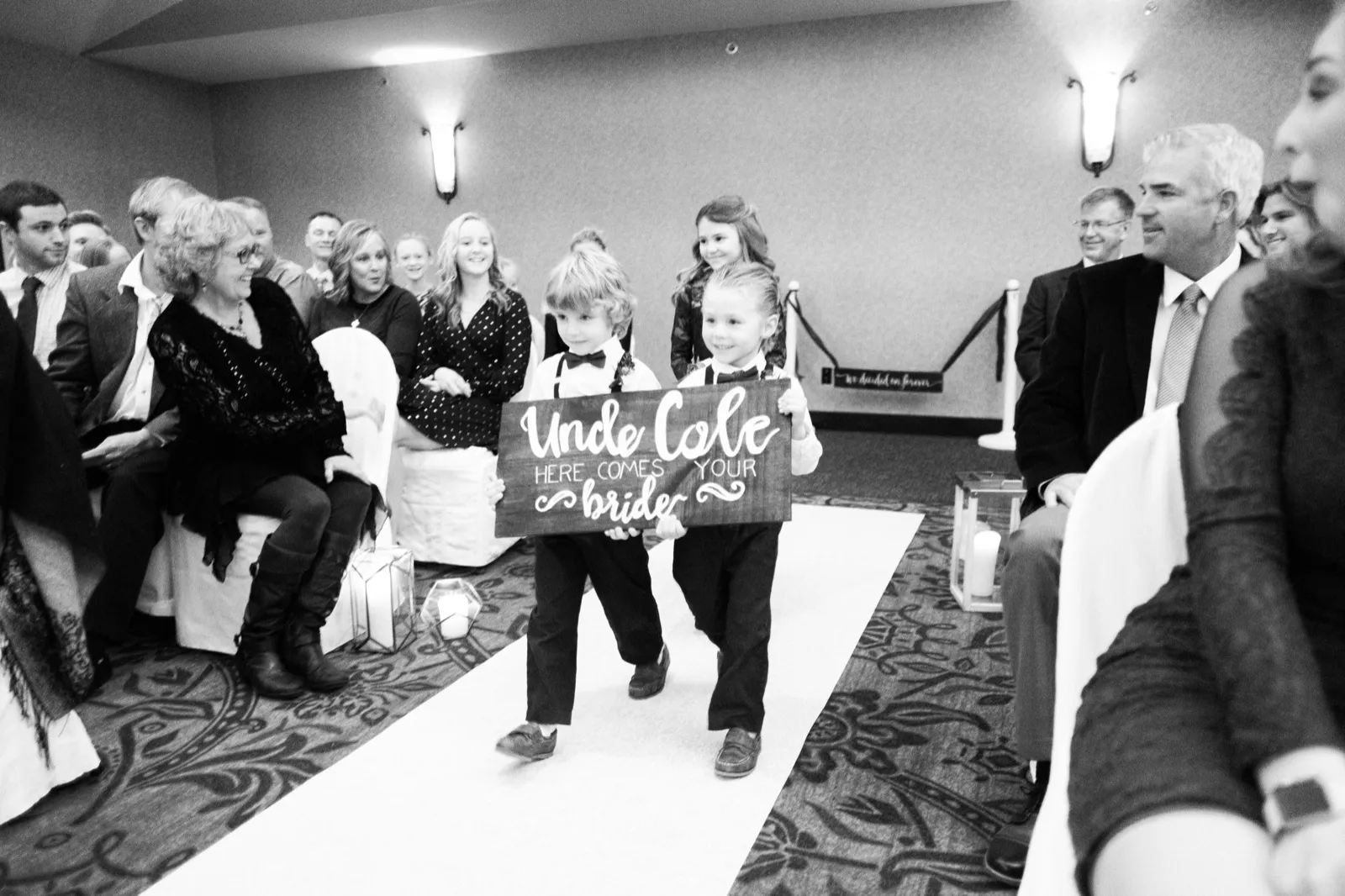 Two young attendants carry a wooden sign reading “Uncle Cole here comes your bride” down the ceremony aisle.