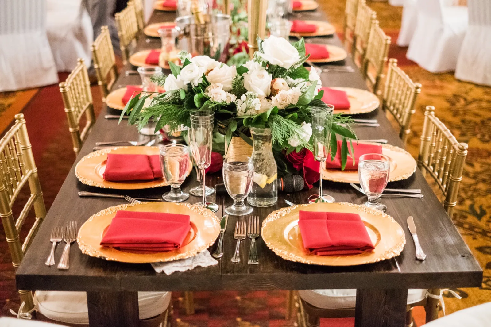 A long reception table is set with gold chargers, red napkins, white flowers, greenery, and gold chairs.
