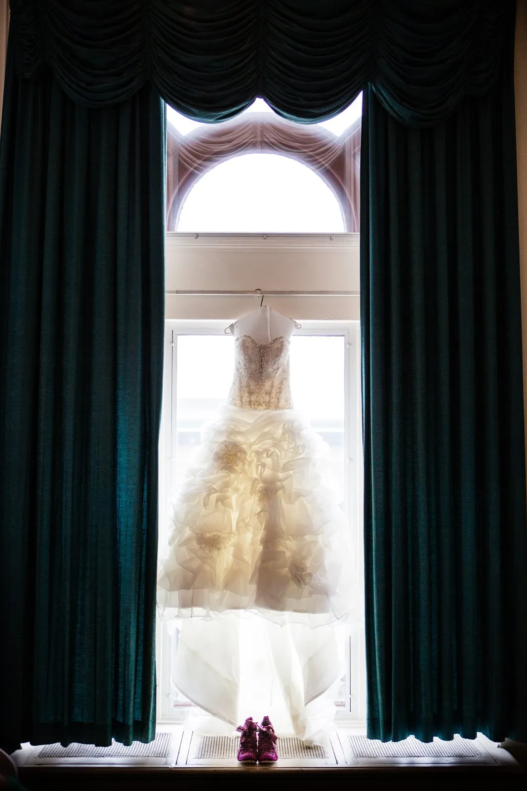 Wedding gown backlit in an arched window framed symmetrically by heavy emerald drapes — Greysolon Plaza bridal suite — Tim Larsen Photography, Duluth MN