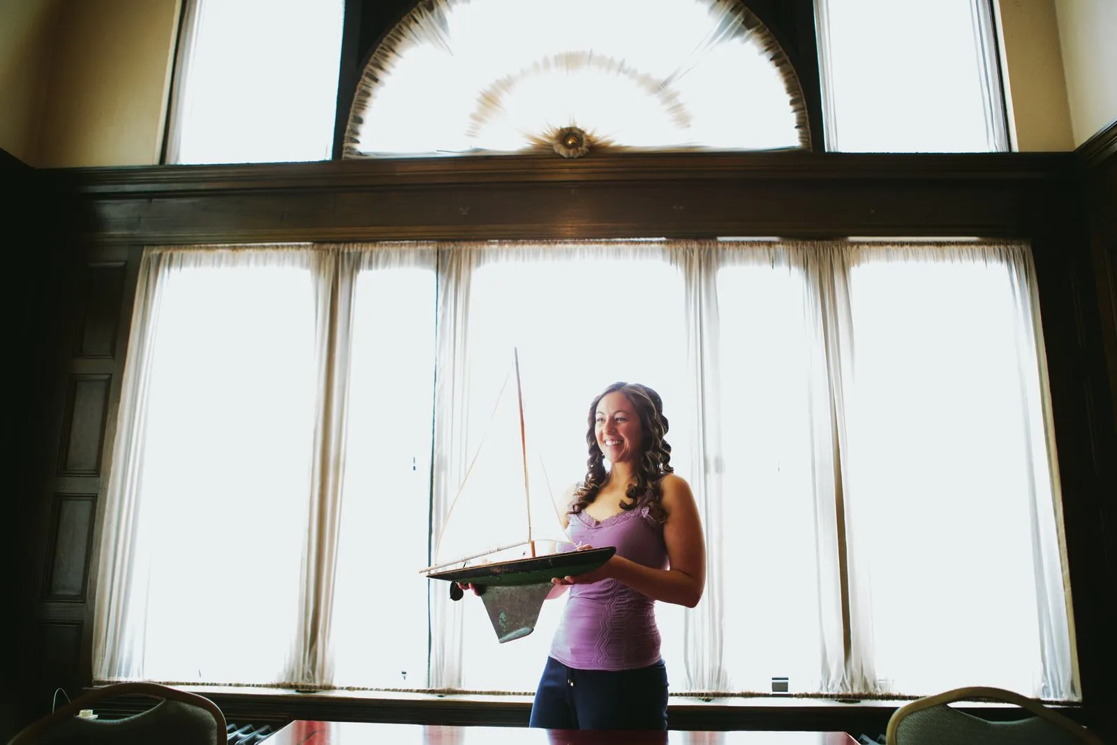 Bride beneath the towering arched windows of the Greysolon Plaza, holding a model sailboat, laughing off-camera — winter wedding — Tim Larsen Photography, Duluth MN