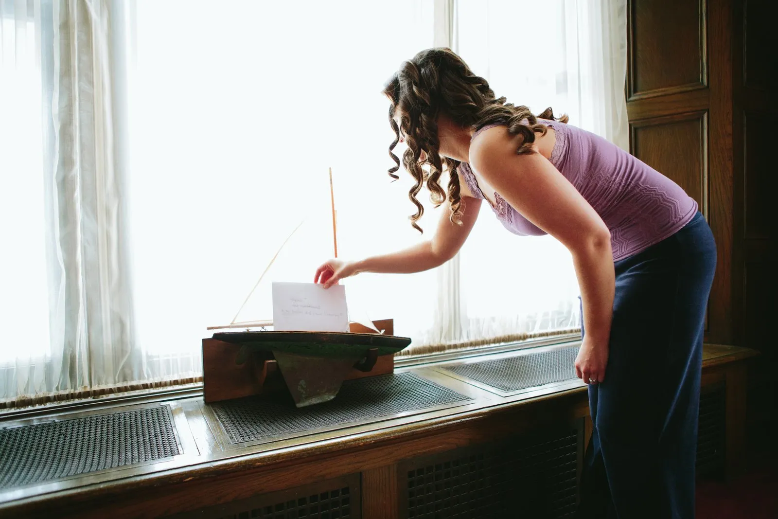 Bride backlit by a tall window reaching for a handwritten note propped on a model sailboat — Greysolon Plaza bridal suite — Tim Larsen Photography, Duluth MN