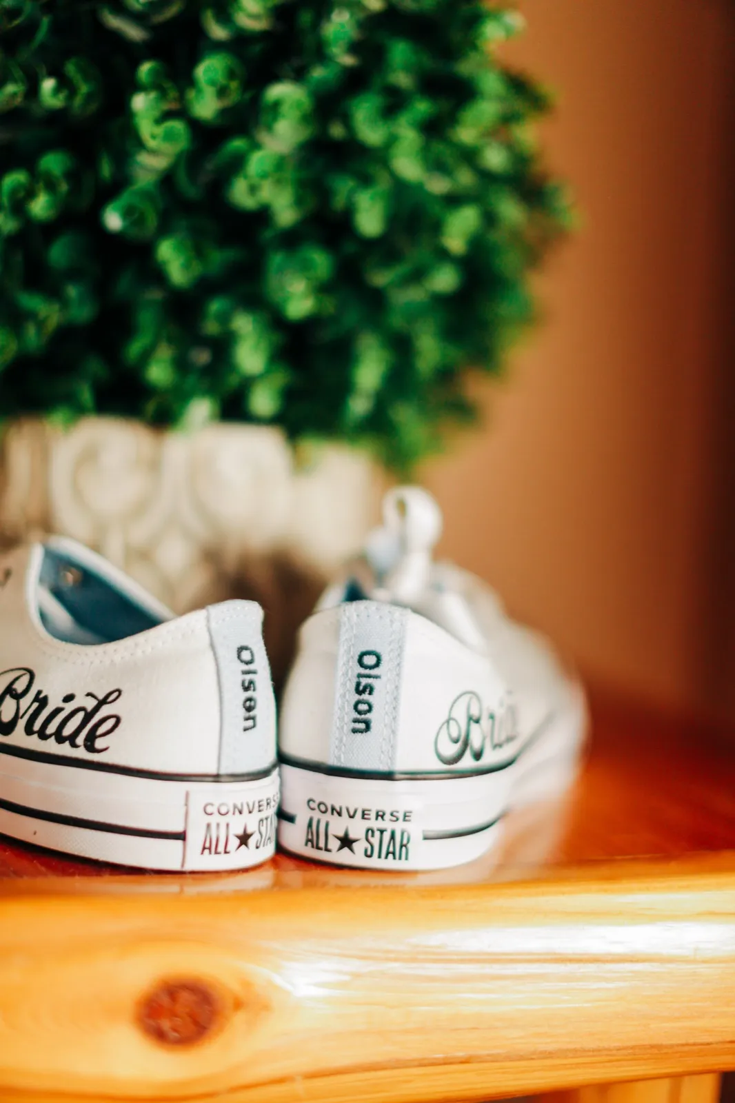 A pair of white Converse All Star sneakers customized with 'Bride' and 'Olson' lettering sit on a wooden surface in front of a green topiary. — Tim Larsen Photography, Brainerd Lakes MN