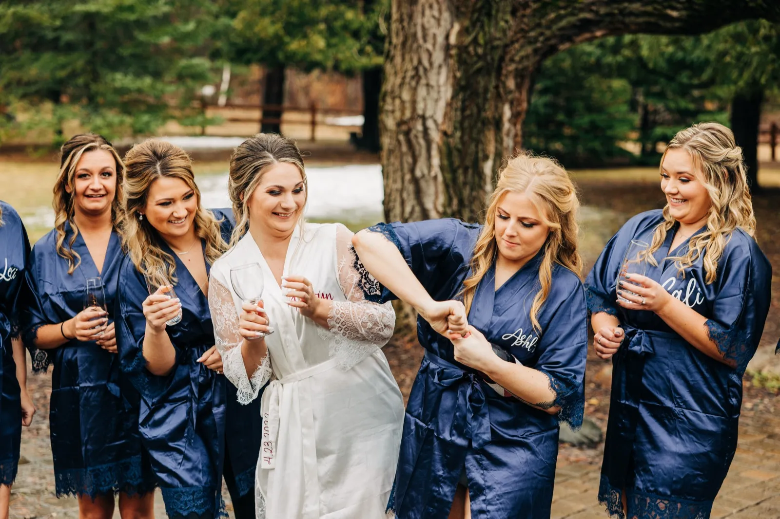 The bride in a white lace robe laughs with four bridesmaids in matching navy robes as one helps adjust her sleeve outdoors among pine trees. — Tim Larsen Photography, Brainerd Lakes MN