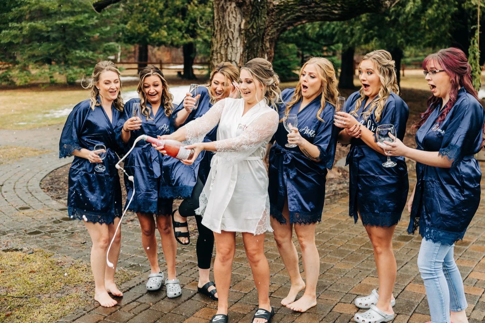 The bride in a white robe sprays champagne while six bridesmaids in matching navy robes react with open-mouthed surprise on a brick path. — Tim Larsen Photography, Brainerd Lakes MN