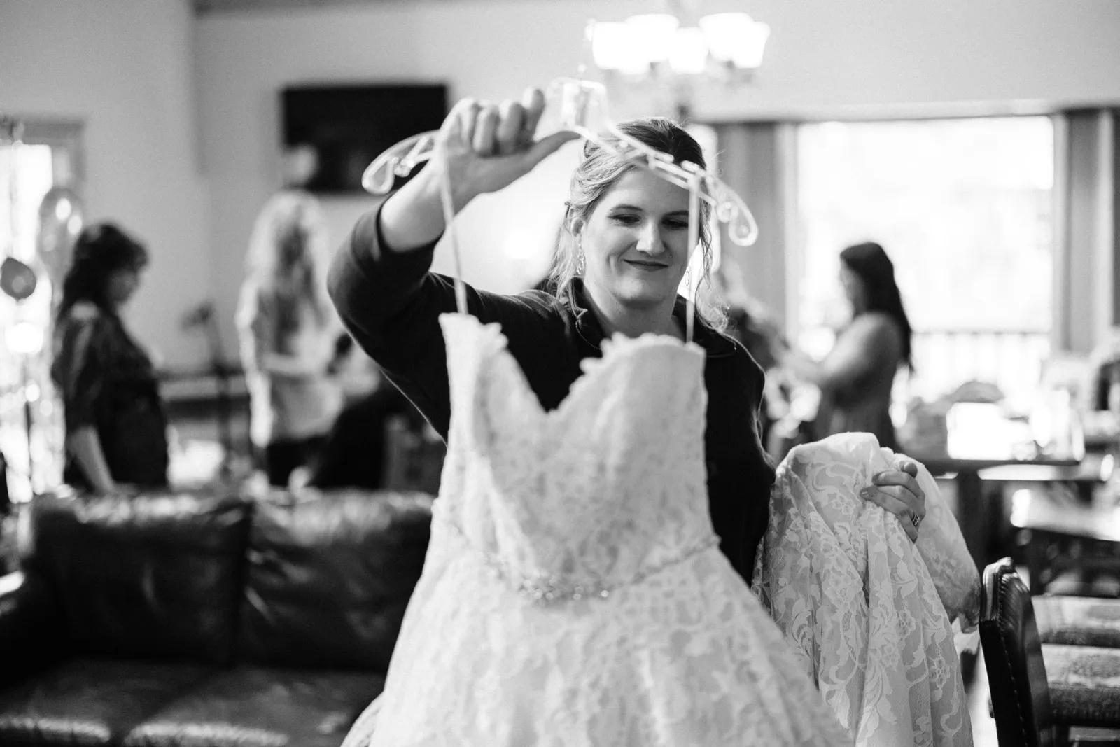 The bride holds her lace wedding dress up by its hangers in a getting-ready room, smiling softly as bridesmaids move in the background. — Tim Larsen Photography, Brainerd Lakes MN