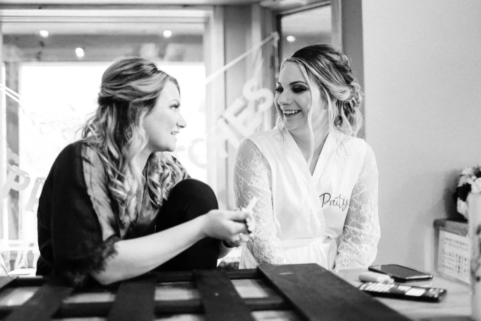 The bride in a lace 'Paity' robe laughs with a bridesmaid across a table in a backlit getting-ready room. — Tim Larsen Photography, Brainerd Lakes MN