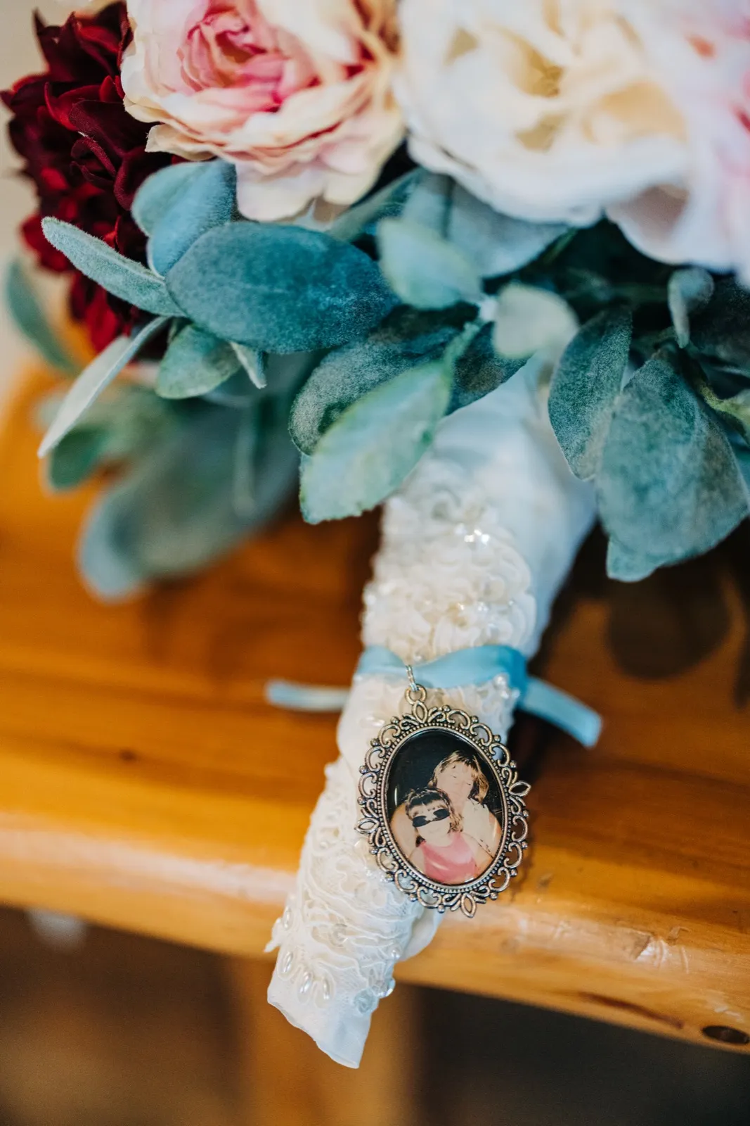 A bridal bouquet of blush, ivory, and deep red roses with lamb's ear leaves rests on a wooden bench, with a small oval memorial locket showing a photograph hanging from a blue ribbon tied around the lace-wrapped stems. — Tim Larsen Photography, Brainerd Lakes MN