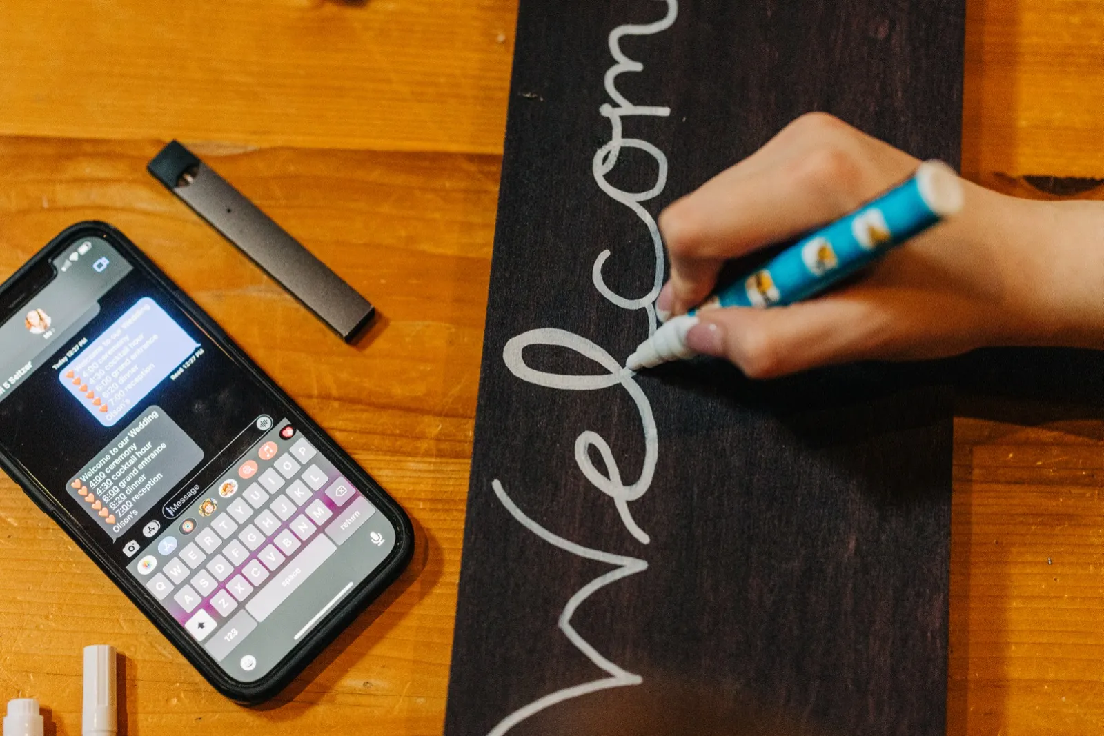 An overhead view shows a hand lettering the word 'Welcome' in white paint marker on a dark wood sign, with a phone displaying the wedding day timeline resting on the table beside it. — Tim Larsen Photography, Brainerd Lakes MN