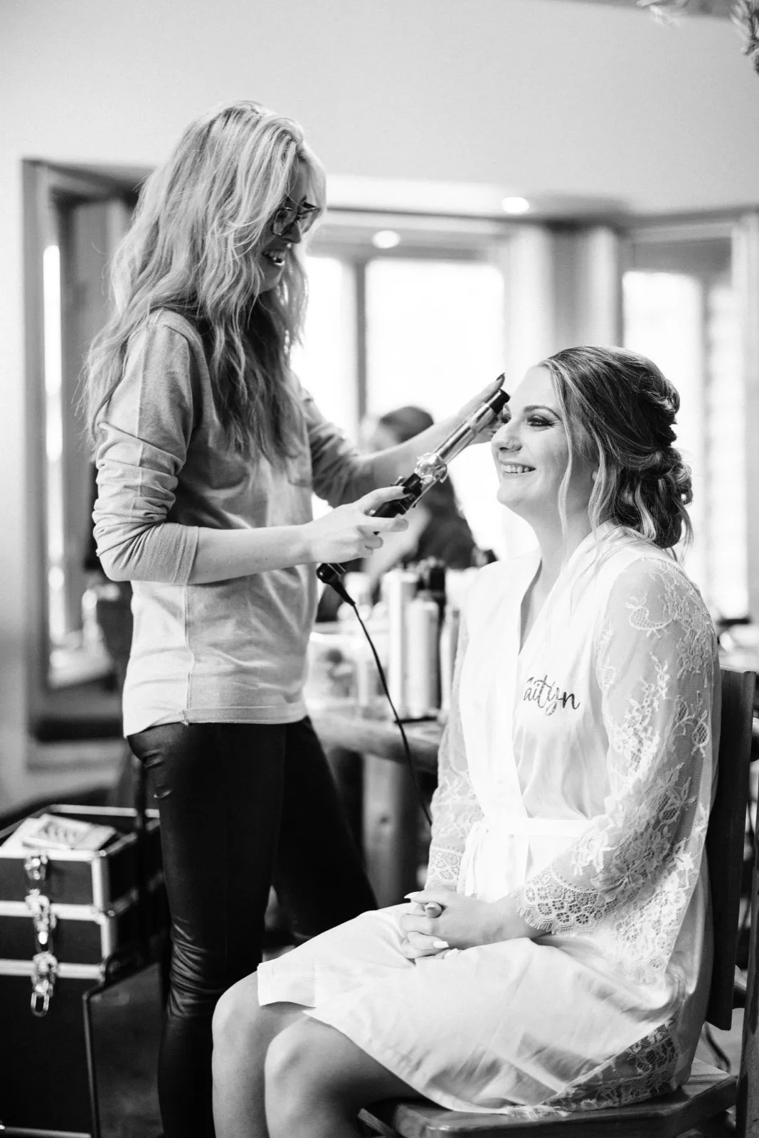 The bride laughs in a lace robe as her stylist curls her hair with a wand in a window-lit bridal suite, rendered in black and white. — Tim Larsen Photography, Brainerd Lakes MN