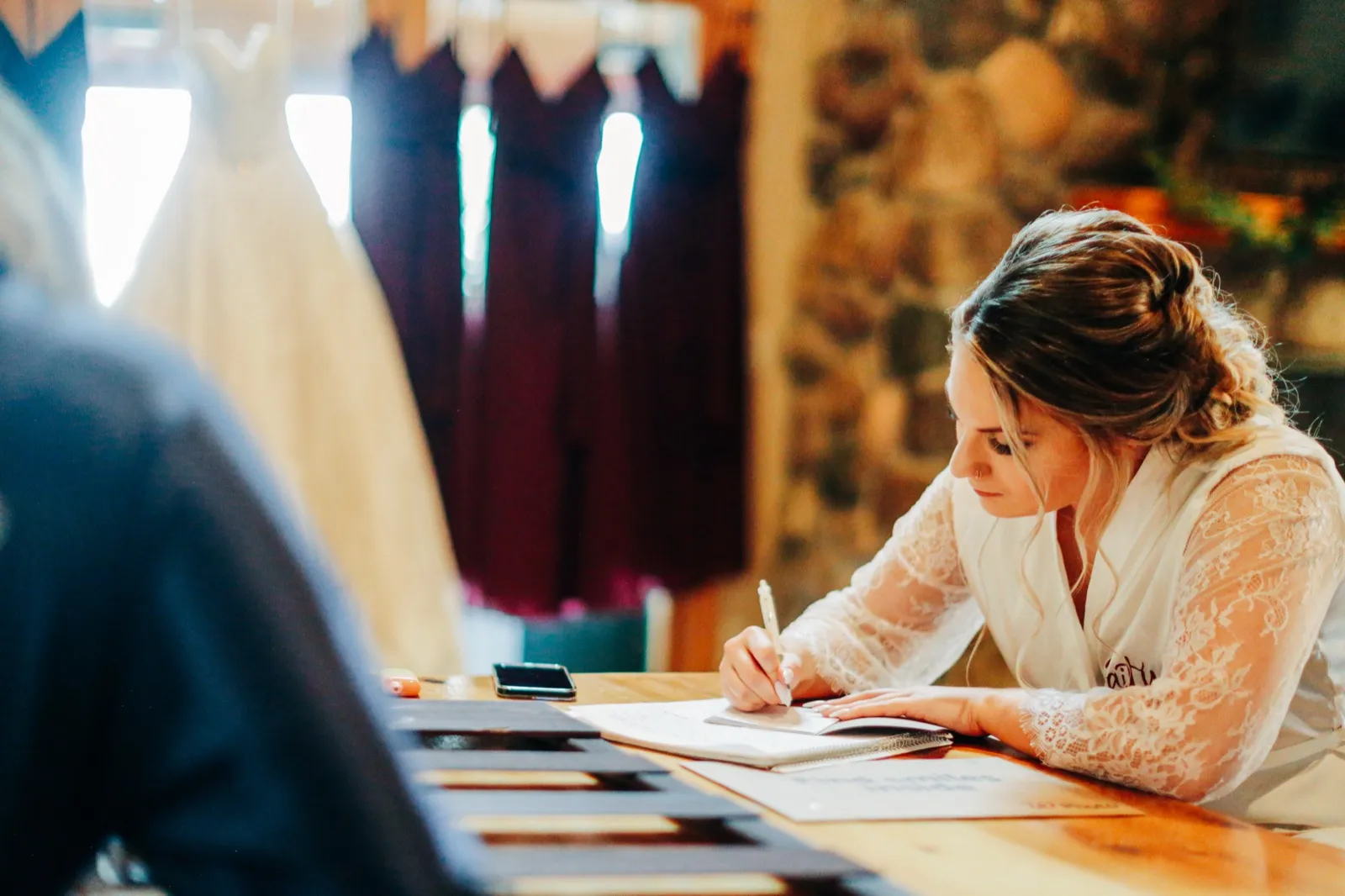 The bride in a lace robe writes a note at a wooden table while her wedding dress and bridesmaid gowns hang in the window light behind her. — Tim Larsen Photography, Brainerd Lakes MN