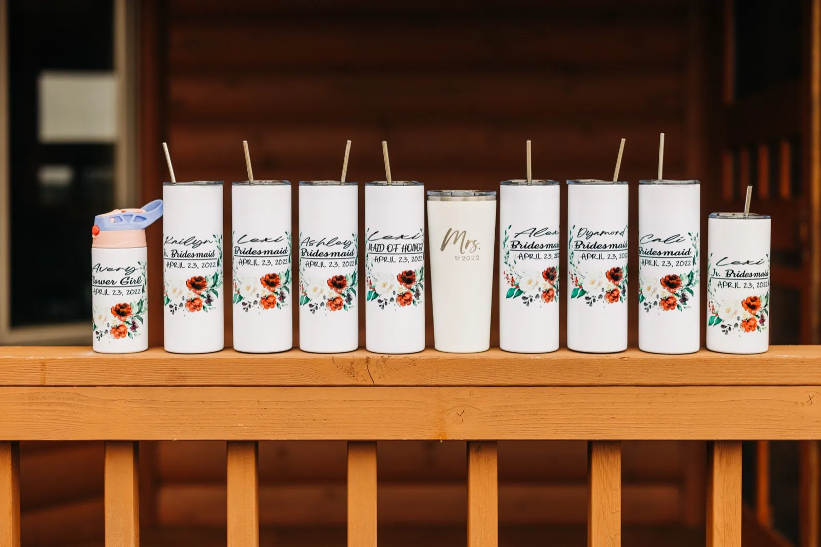 A row of personalized floral tumblers labeled with bridal party roles and the wedding date sits along a wooden porch railing in front of a log cabin. — Tim Larsen Photography, Brainerd Lakes MN